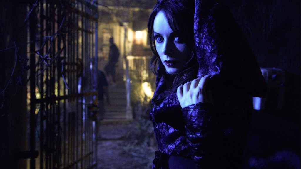 Blind Mag, a woman with artificially enhanced eyeballs, stands ominously staring beside an iron gate